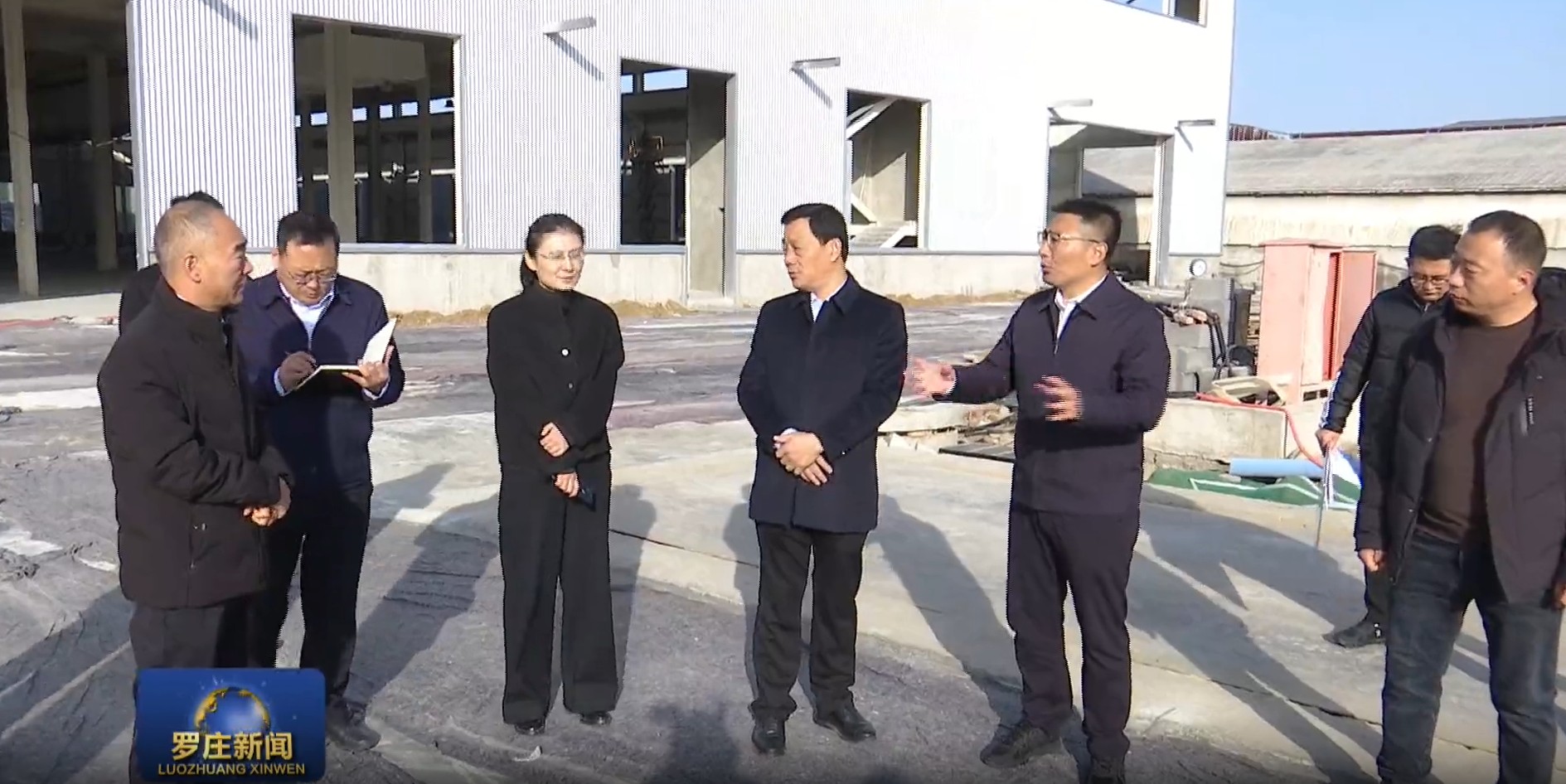 Officials in suits inspect a warehouse construction site outside a large industrial building.